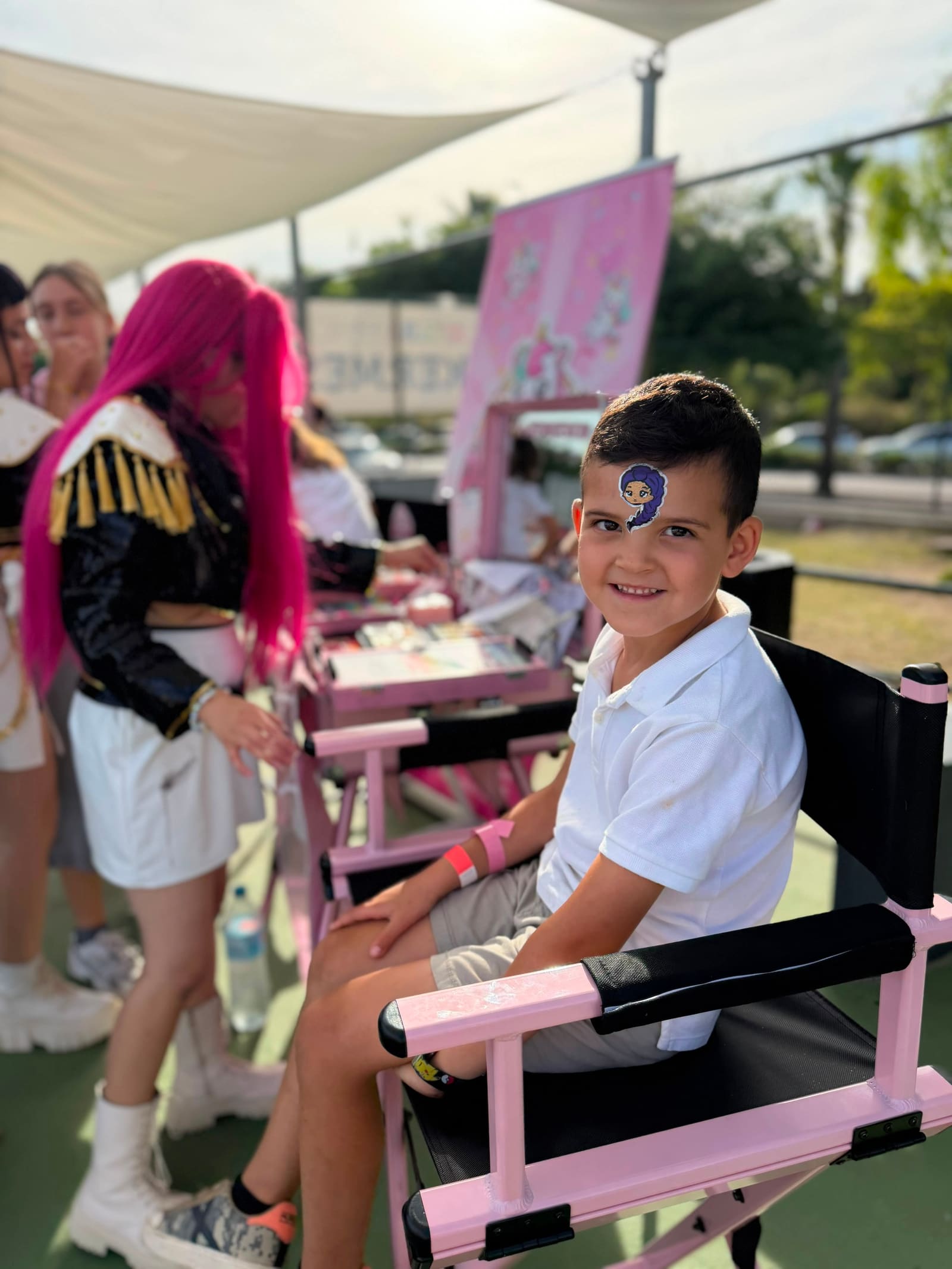 A pink-haired artist does the make-up of a boy sitting in the corner chair