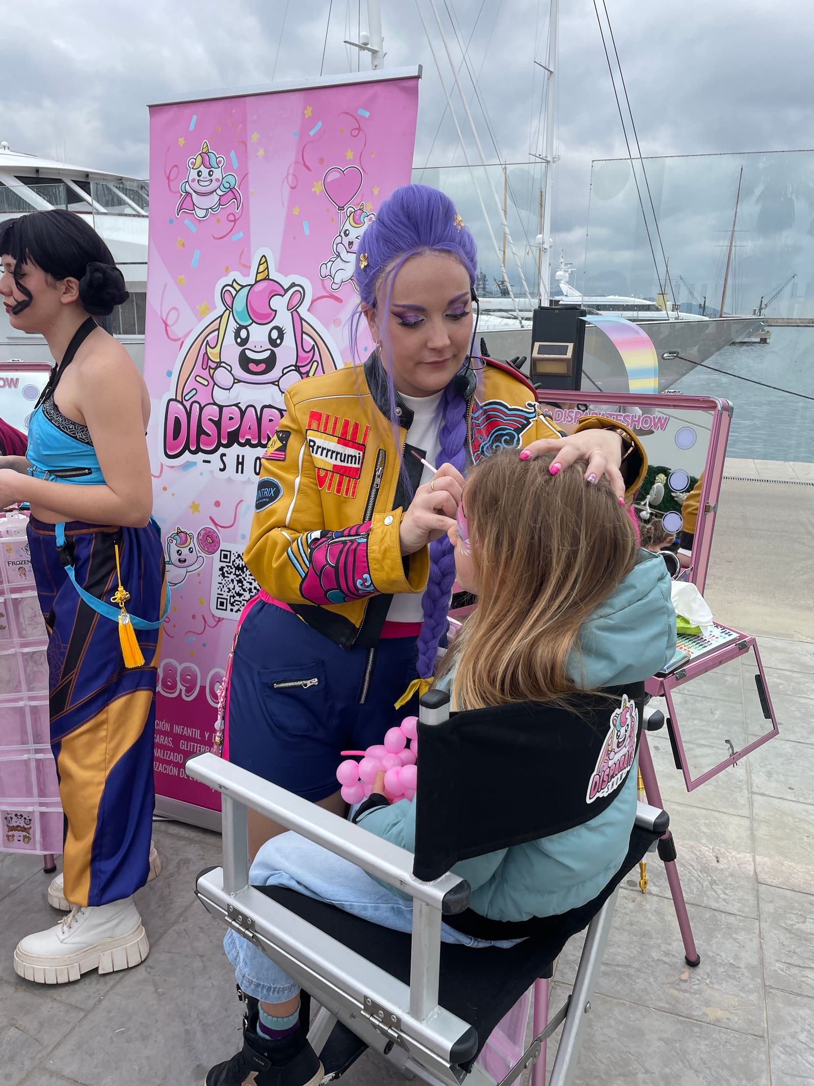 An artist in a yellow jacket does a girl's make-up at the stand, by the harbour