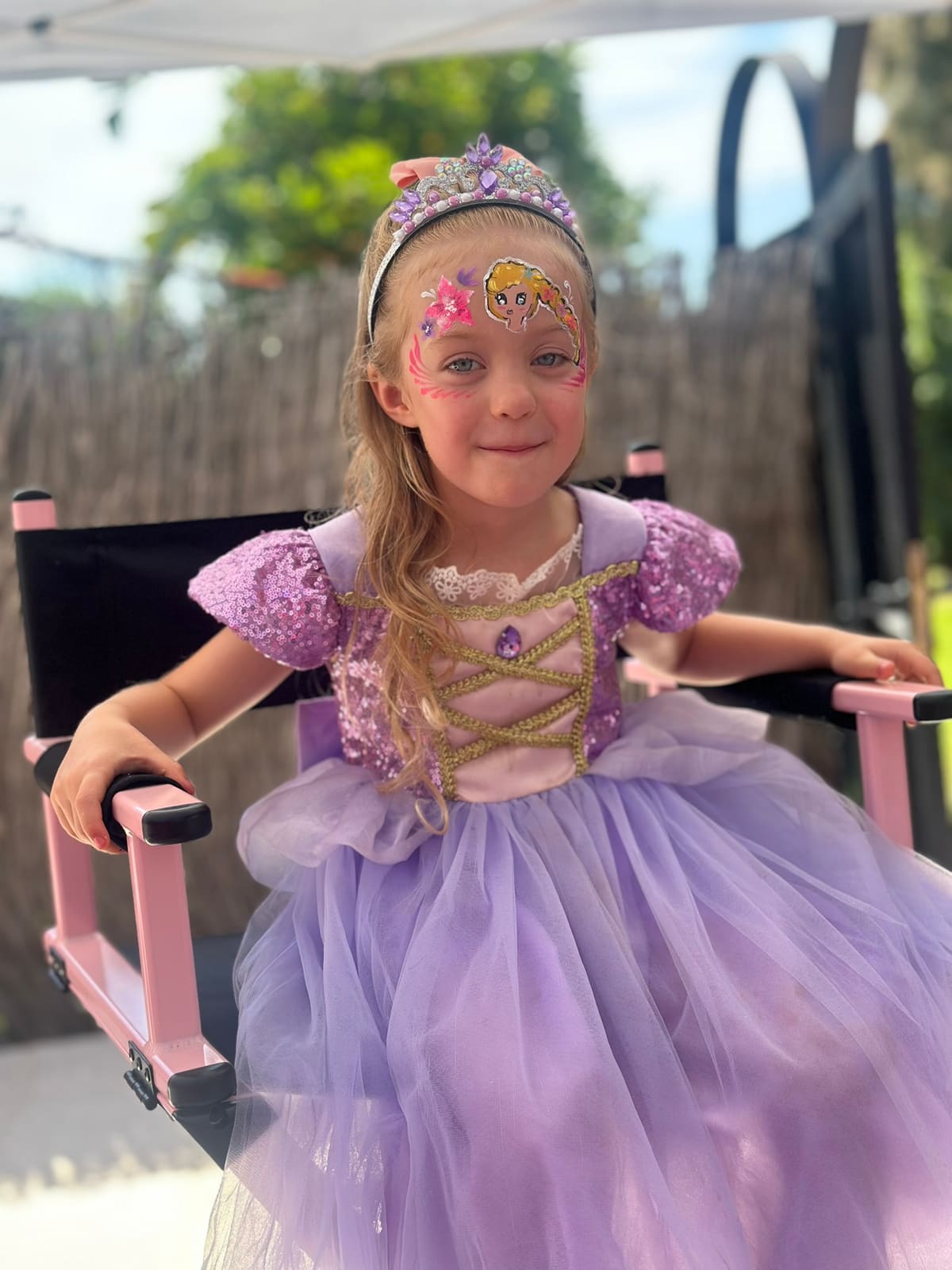 A girl dressed as a princess, wearing a crown, smiles in the make-up chair