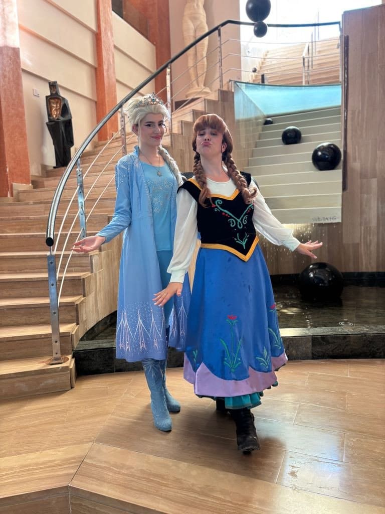 Elsa and Anna, together at the foot of a staircase, open their arms towards the audience