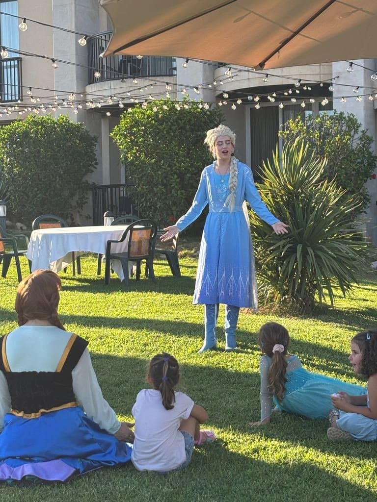 Elsa, alone on the grass with her arms open, and several children sitting on the ground watching her