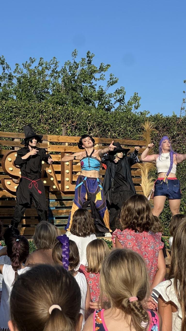 The full K-Pop group on the pallet stage, with a hooded figure to one side