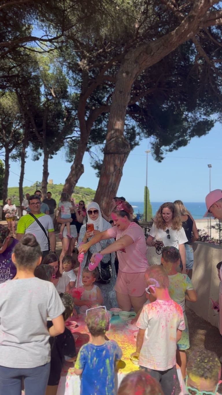 A host pours pink coloured powder from a tub while the children wait their turn, with the sea in the background