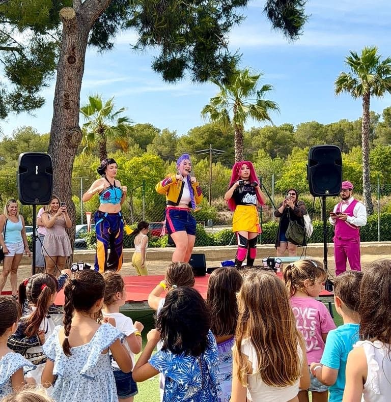 Three K-Pop performers with purple, pink and black hair sing on a stage among palm trees